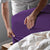 A picture of a man putting a purple fitted sheet over the corner of a bed.