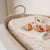 A picture of a moses basket with our Winslow Fitted Sheet on it and a rattle on top of an oatmeal coloured onesie. 